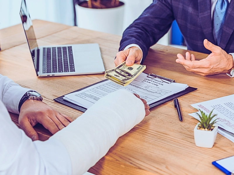 Businessman in blue jacket sitting at table and giving money to worker with broken arm in office.
