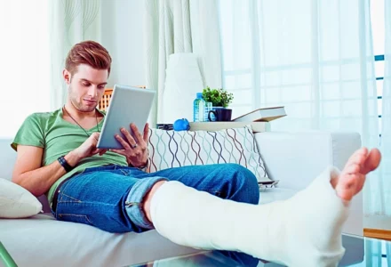 Young man with broken leg sitting on sofa at home and using digital tablet.