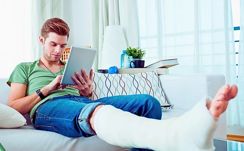 Young man with broken leg sitting on sofa at home and using digital tablet.