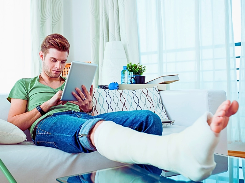 Young man with broken leg sitting on sofa at home and using digital tablet.