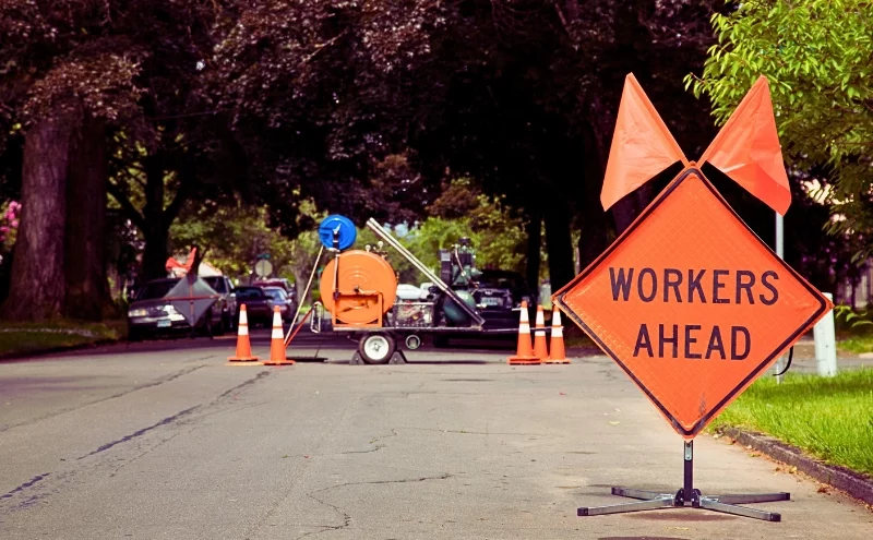 How Road Construction Zones in Naperville Increase Car Accident Risks 1 Orange and Black "Workers Ahead" Sign in Residential Neighbourhood with Equipment in the background. road construction zones