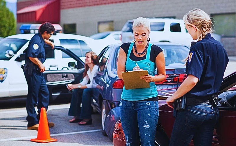 a scene involving a traffic accident with police officers and individuals present. One officer is speaking with a woman sitting on the curb near a police car, while another officer is interacting with a woman standing and holding a clipboard next to a red car. Get Your Naperville Police Crash Report