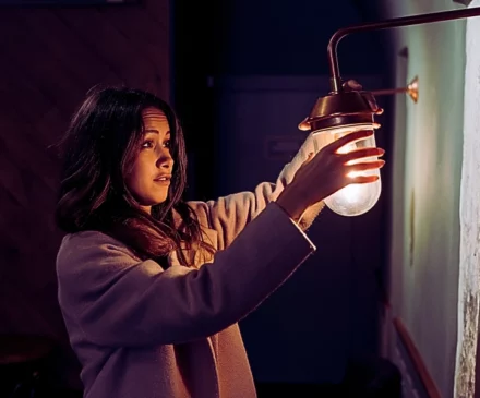 a young woman standing indoors, reaching out with both hands to hold a small wall lamp. Poor Lighting Accident