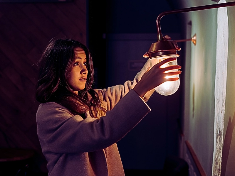 a young woman standing indoors, reaching out with both hands to hold a small wall lamp. Poor Lighting Accident