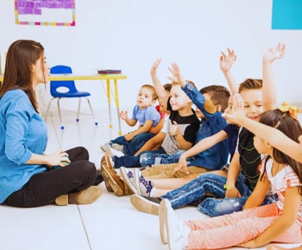 a group of young children sitting on the floor in a classroom setting, all raising their hands as if eager to participate or answer a question. Daycare Injuries