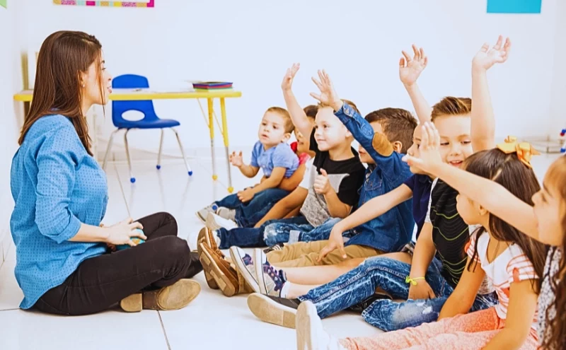 a group of young children sitting on the floor in a classroom setting, all raising their hands as if eager to participate or answer a question. Daycare Injuries
