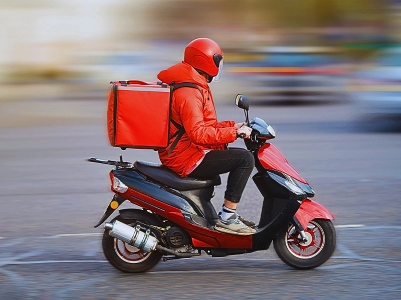a delivery rider wearing a red helmet and jacket, riding a red scooter on a city street. Delivery Driver Accident
