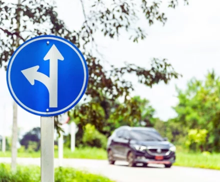 A blue circular traffic sign with two white arrows: one pointing straight ahead and the other curving left. Left-Turn Accidents
