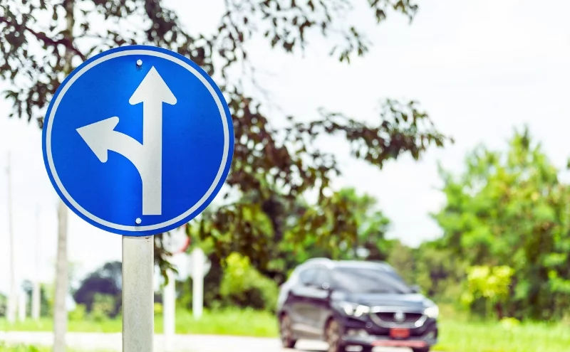 A blue circular traffic sign with two white arrows: one pointing straight ahead and the other curving left. Left-Turn Accidents