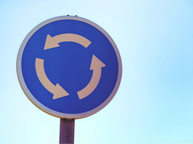 Blue circular traffic sign with white arrows showing roundabout. Crash in a Naperville Roundabout