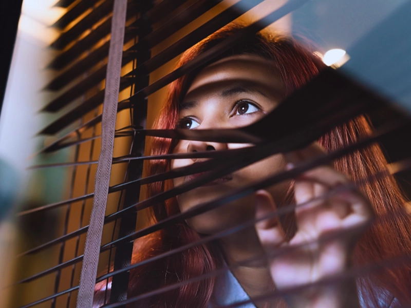 Woman peering curiously through the slats of window blinds. workers' compensation investigators follow you